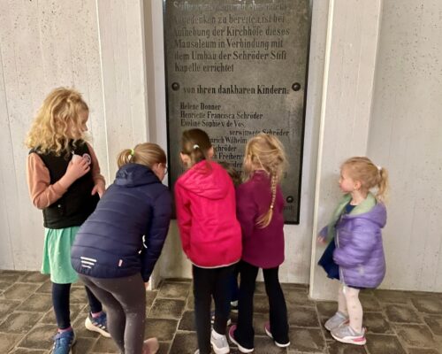 2025_00_29 B1_kk Kinder aus dem gegenüberliegenden Quartier lesen neugierig die Namen der Nachfahren des Stifterpaares, die in der Gruft unter der Kapelle ihre letzte Ruhe gefunden haben.