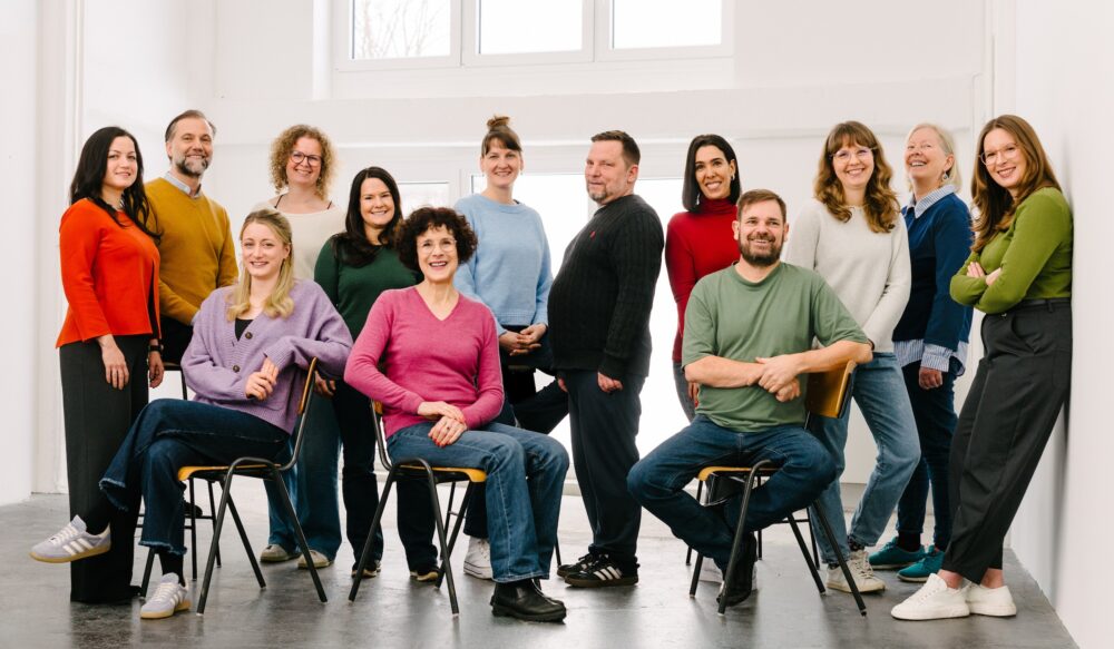 Foto mit 13 Lots*innen in einem lichtdurchfluteten, weißen Raum